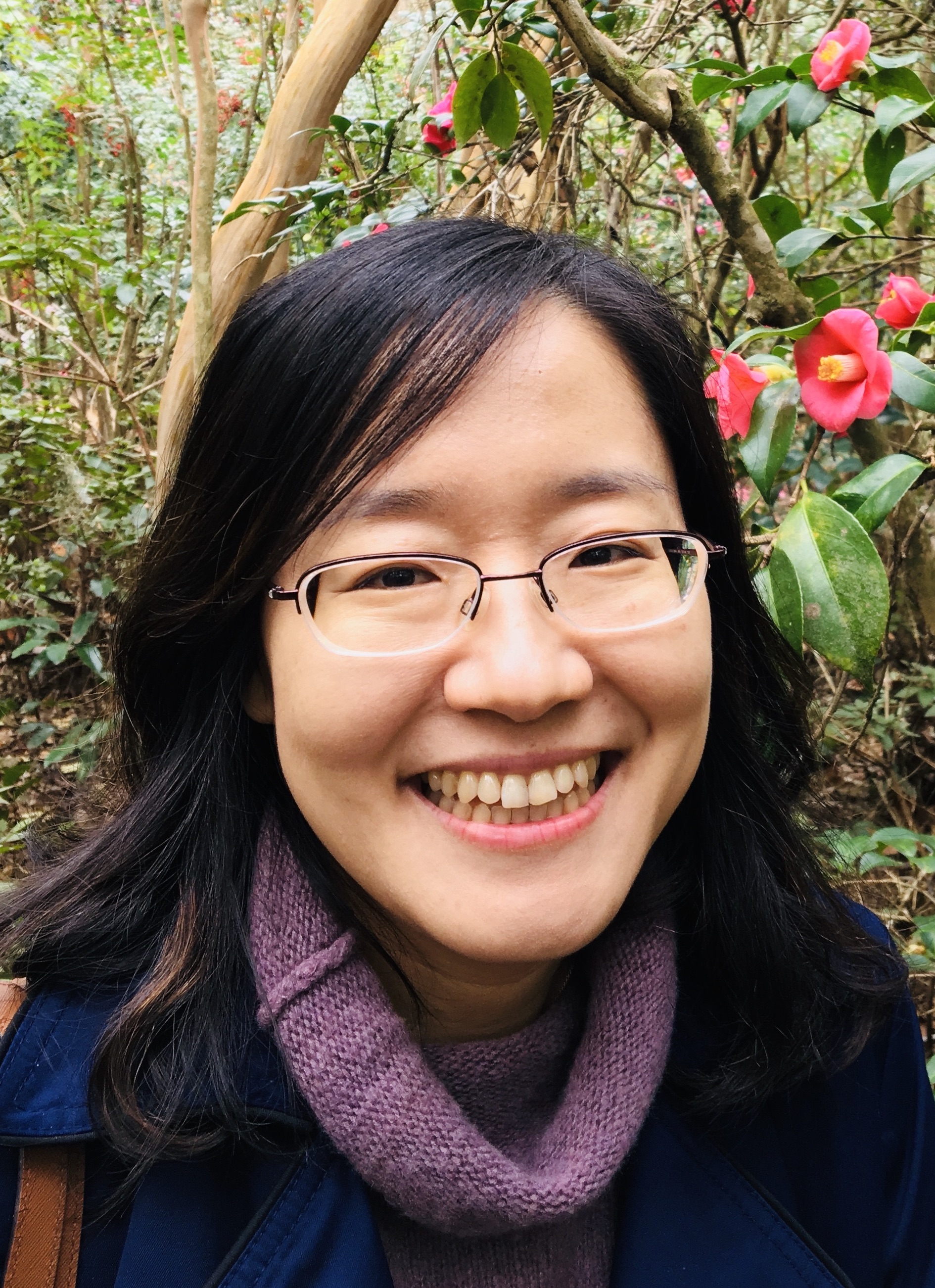Ji Hong has dark, shoulder-length hair and glasses. She is wearing a purple cowl-neck sweater and a blue jacket. She is outside and is surrounded by lush greenery and vibrant red flowers.